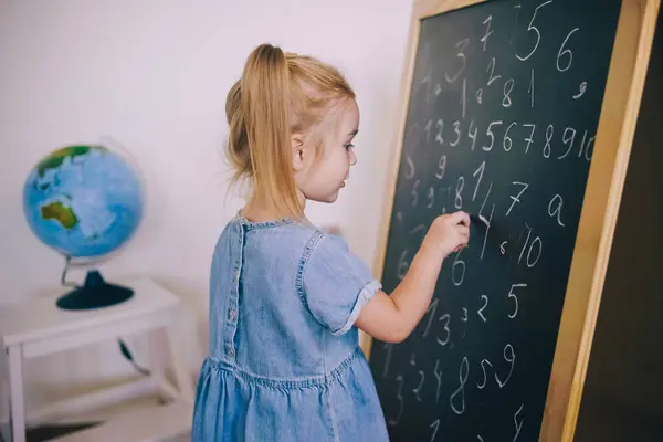 stock image A child is focused on writing numbers on a chalkboard, engaging in a fun learning activity in a cozy room.