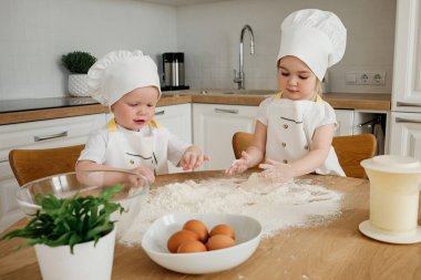İki küçük çocuk, şef şapkası ve önlüğü takıyor, neşeli bir şekilde hamuru parlak mutfaklarında hazırlıyorlar..