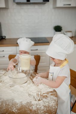 İki çocuk sıcak bir mutfakta, un kaplı bir şekilde yemek pişiriyor. Malzemeleri karıştırıp, mutfak maceralarının tadını çıkarıyorlar..