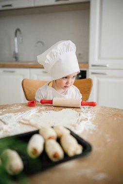 Şefin şapkalı bir çocuk sıcak bir mutfakta malzemelerle çevrili kırmızı bir oklavayla hamuru yuvarlamaya odaklanır..