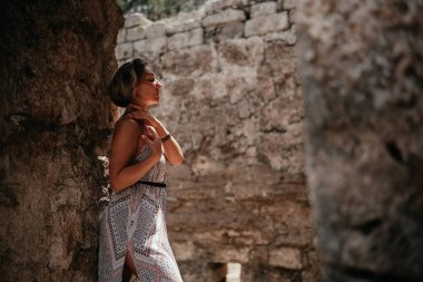 Bir kadın zarif bir şekilde antik taş duvarların arasında duruyor, tarihi kalıntıları keşfederken akışkan desenli elbisesiyle güneşin tadını çıkarıyor..