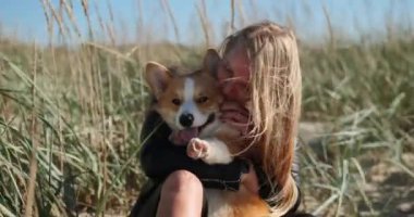 Uzun sarı saçlı bir kadın açık havada Corgi 'yi tutarken gülümsüyor. Köpek de gülümsüyor ve dili dışarı çıkıyor. İkisi de kameraya bakıyor.