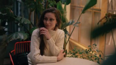 Bir kadın, öğleden sonra rahat bir kafede yemyeşil çiçeklerle çevrili bir masada düşünceli bir şekilde oturur..