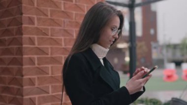 Genç bir kadın akıllı telefonuyla iletişim kuruyor, bir kentin duvarına yaslanırken gülümsüyor..