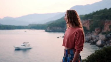 Yaz tatili gezisinde, yaz tatilinde uçurumun kenarındaki kayalıklarda dikilen ve ellerini kaldıran gündelik elbiseli güzel sarışın kadın. Gün batımında resim gibi deniz ve dağ manzarası