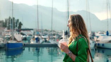 Bir kadın elinde içki tutarken gülümser, gün batımında botlarla dolu sakin bir marinanın yanında durur..