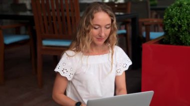 Bir kadın ahşap basamaklarda oturuyor, laptopuna odaklanmış, bir kafede canlı yeşil bitkilerle çevrili..