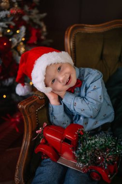 Şenlik şapkası takan genç bir çocuk, güzel süslenmiş bir Noel ağacının yanında oyuncak bir kamyon tutarken tatlı tatlı gülümsüyor..