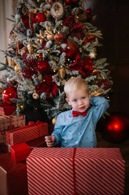 Bir çocuk neşeli bir gülümsemeyle poz verir, etrafı güzel süslenmiş bir ağacın altında hediyelerle çevrilidir..