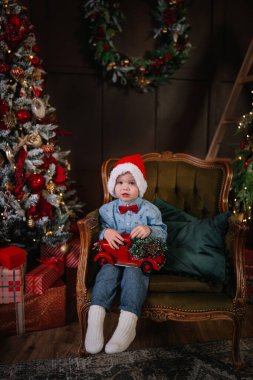 Noel Baba şapkası takan küçük bir çocuk, Noel ağacının yanında oyuncak bir kamyon tutuyor..