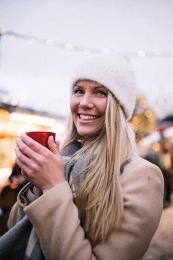 Neşeli genç bir kadın sıcak bir içki tutuyor, açık hava kış pazarında neşeyle gülümsüyor..