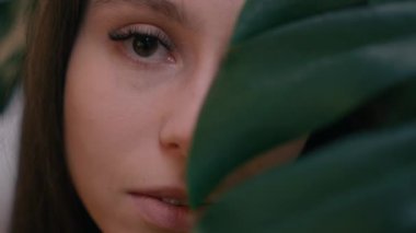 Gözlüklü genç bir kadın, dingin ve doğal bir ortamda yemyeşil tropikal yaprakların arkasından dışarı bakar..