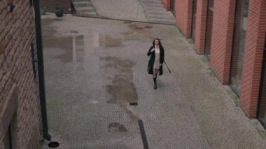 Neşeli bir kadın bej bir elbise ve siyah bir palto giyiyor. Çağdaş bir yapının yakınındaki kaldırım taşı yolda yürüyor..