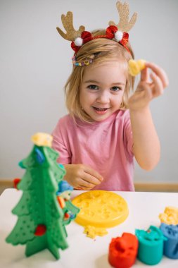 Ren geyiği boynuzları olan neşeli bir çocuk renkli mankenlik hamuru kullanarak tatil süslemeleri yapıyor..