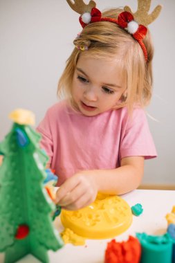 Ren geyiği boynuzları olan neşeli bir çocuk renkli mankenlik hamuru kullanarak tatil süslemeleri yapıyor..
