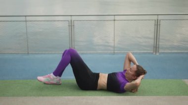 Bir kadın açık havada su kenarında egzersiz yapıyor, spor kıyafetleriyle spor minderinde mekik çekiyor..