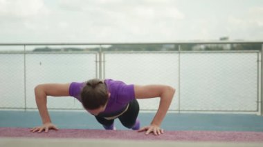 Bir kadın bulutlu gökyüzünün altındaki bir parkta şınav çekerek egzersiz yapıyor ve spor rutinine odaklanıyor..