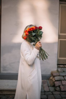 Beyaz önlüklü bir kadın kaldırım taşlı bir sokakta duruyor, yüzüne yakın renkli çiçekler tutuyor, anın tadını çıkarıyor..