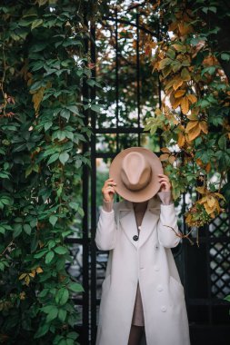 Bir insan canlı yeşil bitkiler ve güz yapraklarıyla çevrili bir bahçede elinde kahverengi bir şapkayla duruyor..