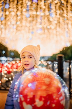 Kışlık ceketli neşeli bir çocuk elinde parlak bir balonla şenlikli bir etkinlikte göz kamaştırıcı ışıkların önünde duruyor..