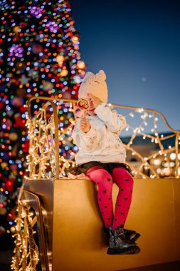 Sıcak şapkalı ve pembe taytlı bir çocuk lolipopu olan bir kızağın üzerinde oturuyor, etrafı şenlikli ışıklar ve Noel ağacı ile çevrili..