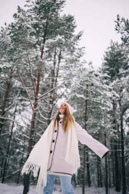 Aktif, beyaz yün ceketli, şapkalı ve uçan atkılı güzel bir kadın karlı kış ormanının tadını çıkarıyor.