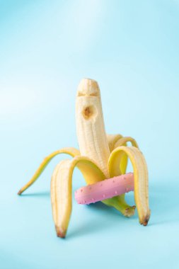 Bir muz dikey durur, kısmen soyulmuş, kabuğunda pembe bir çörek, açık mavi bir arka planda..