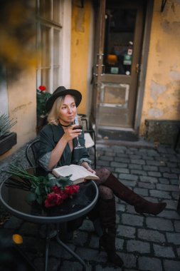 Çiçeklerle çevrili tuhaf bir masada kitap okurken bir kadın içkisini yudumluyor..