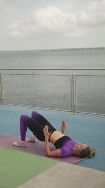 Bir kadın su kenarında hasırda egzersiz yapıyor, güneş ışığında tek bacaklı kalça köprüsü yapıyor..