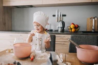 Sevimli gülümseyen 3 yaşındaki aşçı şapkalı ve önlüklü kız una üflüyor, ev mutfağında yemek pişiriyor..