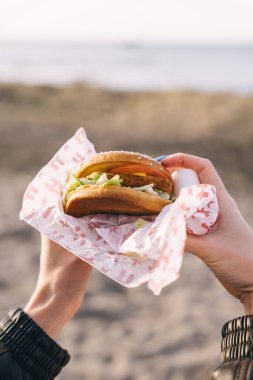 Tallinn, Estonya - 2 Nisan 2025: Bir insan gün batımında sahilde durup anın tadını çıkarırken kağıda sarılı lezzetli bir burger tutar.