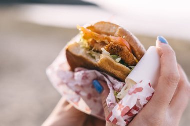 Tallinn, Estonya - 2 Nisan 2025: Bir kişi arka planda plaj ve gün batımı olan kağıda sarılı bir burger tutuyor.