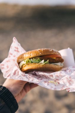 Tallinn, Estonya - 2 Nisan 2025: Bir el, güneşli bir günde plajdaki katmanlarını gösteren kağıda sarılı taze Hesburger 'i sunar..