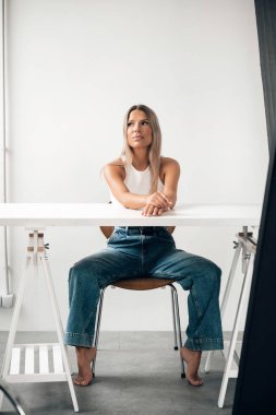 Genç bir kadın minimalist bir masada rahat bir şekilde oturuyor, parlak bir ortamda kendine güvenen bir ifade sergiliyor..