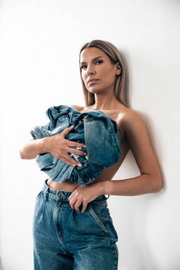 Modern bir stüdyo ortamında beyaz bir duvara karşı poz veren bir model kot elbise sergiliyor..