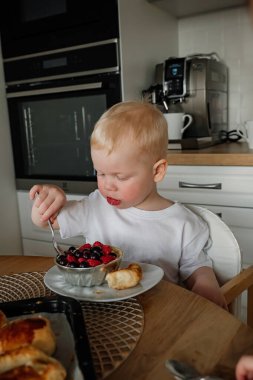 Yeni yürümeye başlayan bir çocuk, çağdaş bir mutfak ortamında kruvasanın yanında bir kase taze karışık böğürtlen yer..