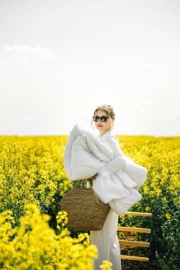 Güneş gözlüklü bir kadın açık gökyüzünün altında canlı sarı bir çiçek tarlasında dururken rahat bir battaniye tutuyor..
