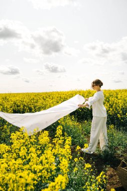 Beyaz elbiseli bir kadın, açık bir gökyüzünün altındaki sarı bir çiçek tarlasına kumaş serer..