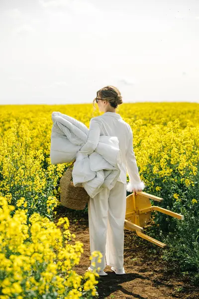 Sarı çiçeklerle dolu bir tarlada yürüyen bir kadın, güneşli günün tadını çıkarırken battaniyeler ve küçük bir sandalye taşıyor..