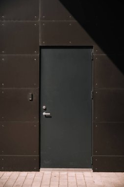 Pürüzsüz, karanlık bir kapı gölgeli bir duvarın karşısında duruyor, öğleden sonra ışığında ilgi çekici bir kontrastlık yaratıyor..