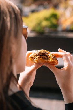 Uzun saçlı bir kadın dışarıda, parlak güneş ışığında otururken çikolatalı kurabiyenin tadını çıkarıyor..