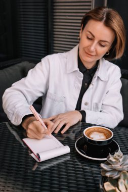 Bir kadın modern bir kafe masasında oturuyor, not defterine notlar yazıyor, yanında bir fincan kahveyle..