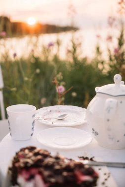 Güneş çiçekli bitkilerin arkasında batarken göl kenarındaki bir masada çay ve tatlının tadını çıkararak sakin bir atmosfer yaratıyor..
