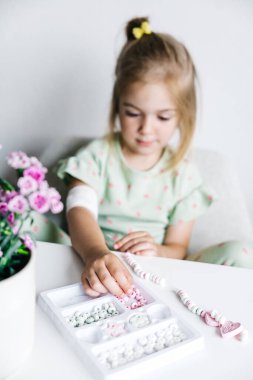 Bir kız, çiçeklerle ve renkli boncuklarla çevrili bir masada boncuklu bir tasarım yapmaya odaklanır..