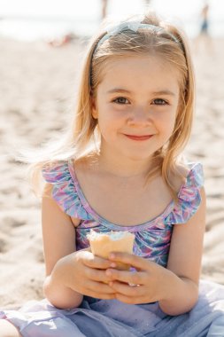 Gülümseyen kız kumlu sahilde otururken elinde dondurma külahıyla güneşli bir günün tadını çıkarıyor..