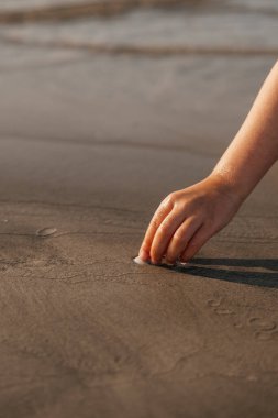 Gün batımında dalgalar yavaşça kıyıya vururken bir çocuk kumsaldaki ıslak kuma dokunmak için uzanır..
