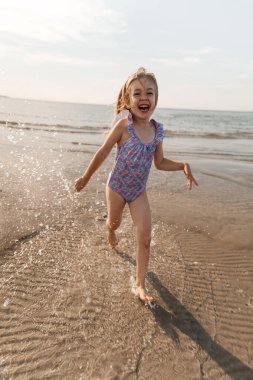 Bir kız gün batımında sahildeki sığ dalgalarda neşeyle koşar, saf mutluluğu gösterir..