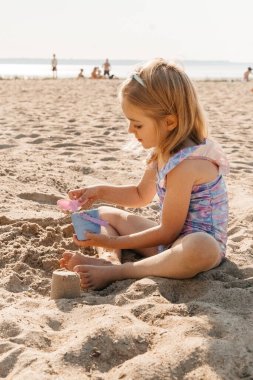 Genç bir kız kumsalda oynar, kumdan kale yapar ve güneşli bir günün tadını çıkarır..