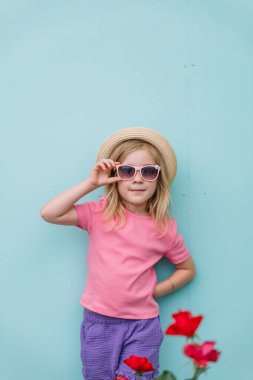 Güneş gözlüğü takan ve şapka takan kız kendine güvenen bir duvar ve güzel çiçeklerin yanında duruyor..