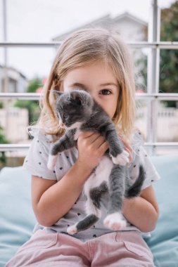 Genç bir kız, elinde neşeli, gri ve beyaz bir kedi yavrusunu tutarken gülümsüyor..